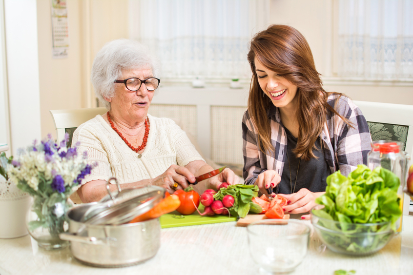 Alte Frau gemeinsam mit junger Frau an Tisch sitzend und schneiden Tomaten. 
